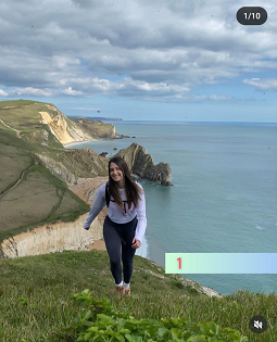 A white female with long brown hair smiles at the camera on a coastal path. Durdle Door is in the background. 